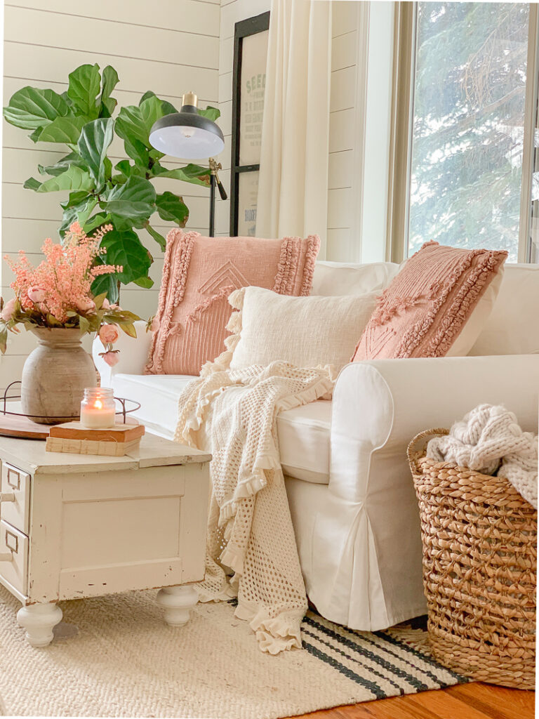 A photo of a sofa with three pillows in white and pink, perfect for spring, accompanied by a plant, a basket, and a table.